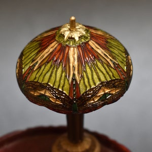 Pode incluir: Uma l&acirc;mpada de vitral em miniatura com um design verde, vermelho e dourado. A l&acirc;mpada est&aacute; sobre uma base de madeira.
