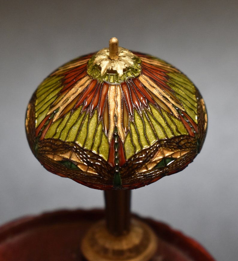 Pode incluir: Uma l&acirc;mpada de vitral em miniatura com um design verde, vermelho e dourado. A l&acirc;mpada est&aacute; sobre uma base de madeira.