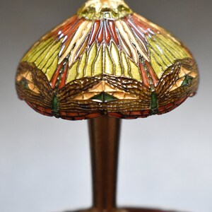 Pode incluir: Uma l&acirc;mpada de mesa em miniatura de estilo Tiffany com um abajur de vidro colorido que apresenta um design de lib&eacute;lula em tons de verde, vermelho e dourado. A l&acirc;mpada tem uma base de bronze e est&aacute; sobre uma mesa de madeira.