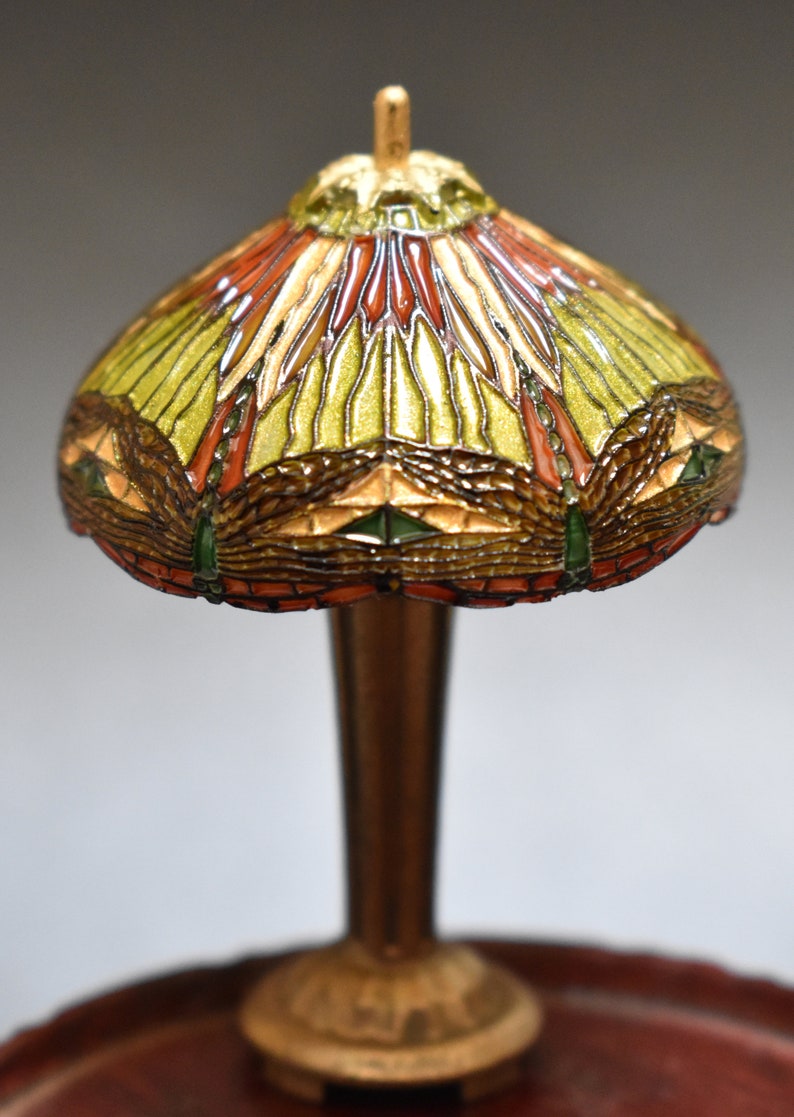 Pode incluir: Uma l&acirc;mpada de mesa em miniatura de estilo Tiffany com um abajur de vidro colorido que apresenta um design de lib&eacute;lula em tons de verde, vermelho e dourado. A l&acirc;mpada tem uma base de bronze e est&aacute; sobre uma mesa de madeira.