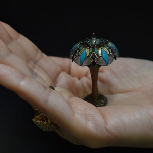 K&ouml;nnte beinhalten: Eine Tiffany-Lampe im Miniaturformat mit einem t&uuml;rkisfarbenen und goldenen Buntglas-Schirm. Die Lampe steht in der Handfl&auml;che.