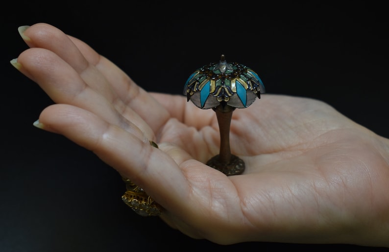 K&ouml;nnte beinhalten: Eine Tiffany-Lampe im Miniaturformat mit einem t&uuml;rkisfarbenen und goldenen Buntglas-Schirm. Die Lampe steht in der Handfl&auml;che.
