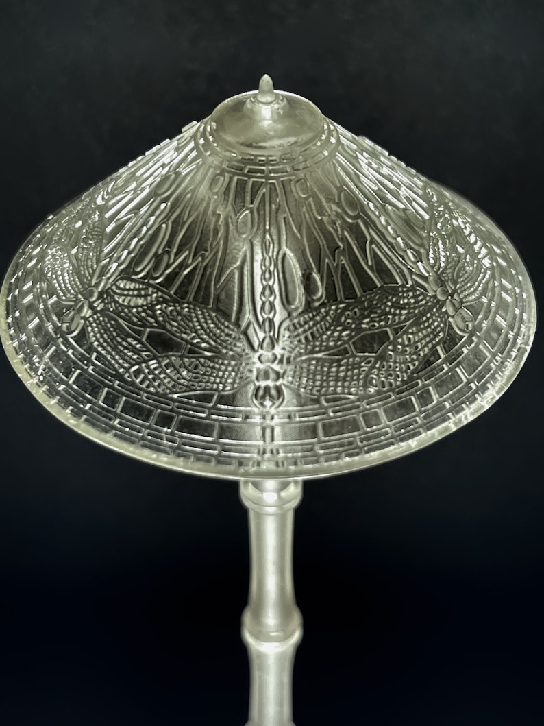 Op de afbeelding: Een heldere glazen tafellamp met een libellenontwerp op de kap. De lamp heeft een zilveren voet en een witte kap.