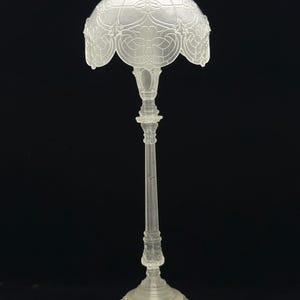 May include: A clear, ornate lamp with a detailed floral design on the shade. The lamp has a tall, slender stem and a decorative base. The shade is approximately 6 inches in diameter.