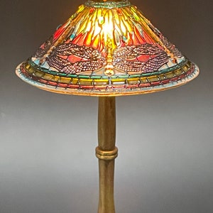 Op de afbeelding: Een gouden tafellamp met een gebrandschilderd lampenkap met een libellenmotief. De kap is gemaakt van gekleurde glasstukken in tinten rood, groen, blauw en geel.