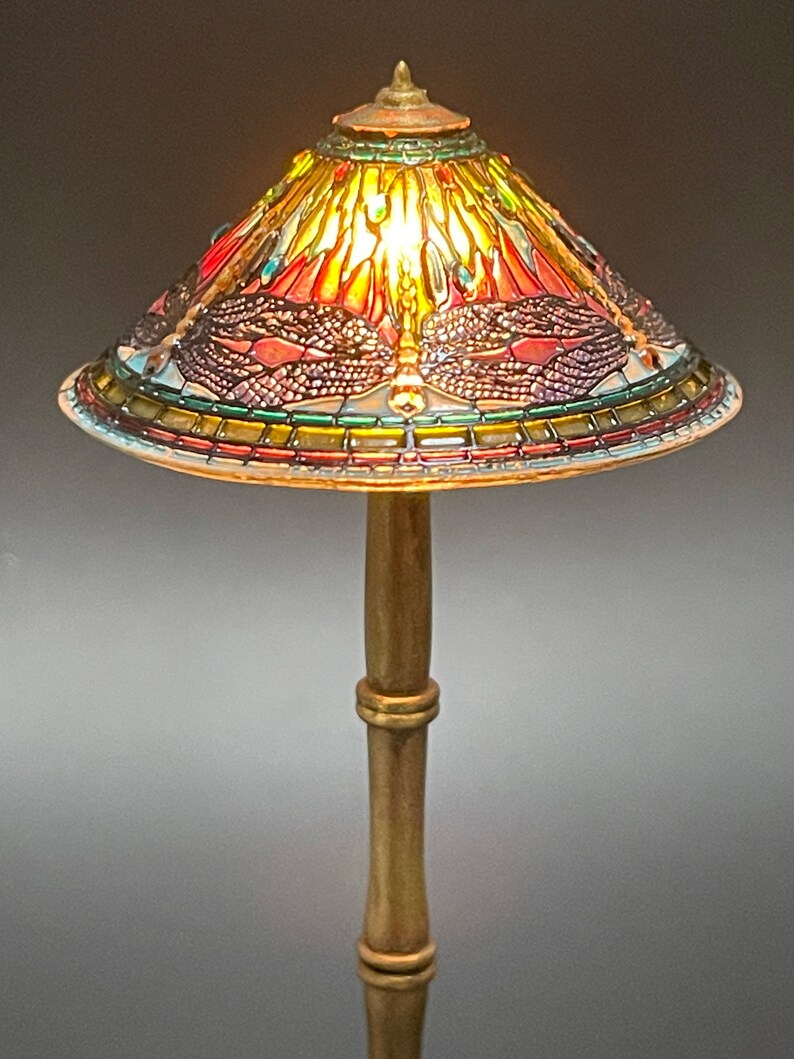 Op de afbeelding: Een gouden tafellamp met een gebrandschilderd lampenkap met een libellenmotief. De kap is gemaakt van gekleurde glasstukken in tinten rood, groen, blauw en geel.