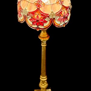 May include: A decorative floor lamp with a stained glass shade. The shade features a floral design in shades of red, orange, and yellow. The base and stem are gold-colored, with ornate detailing. The lamp is a vintage style.
