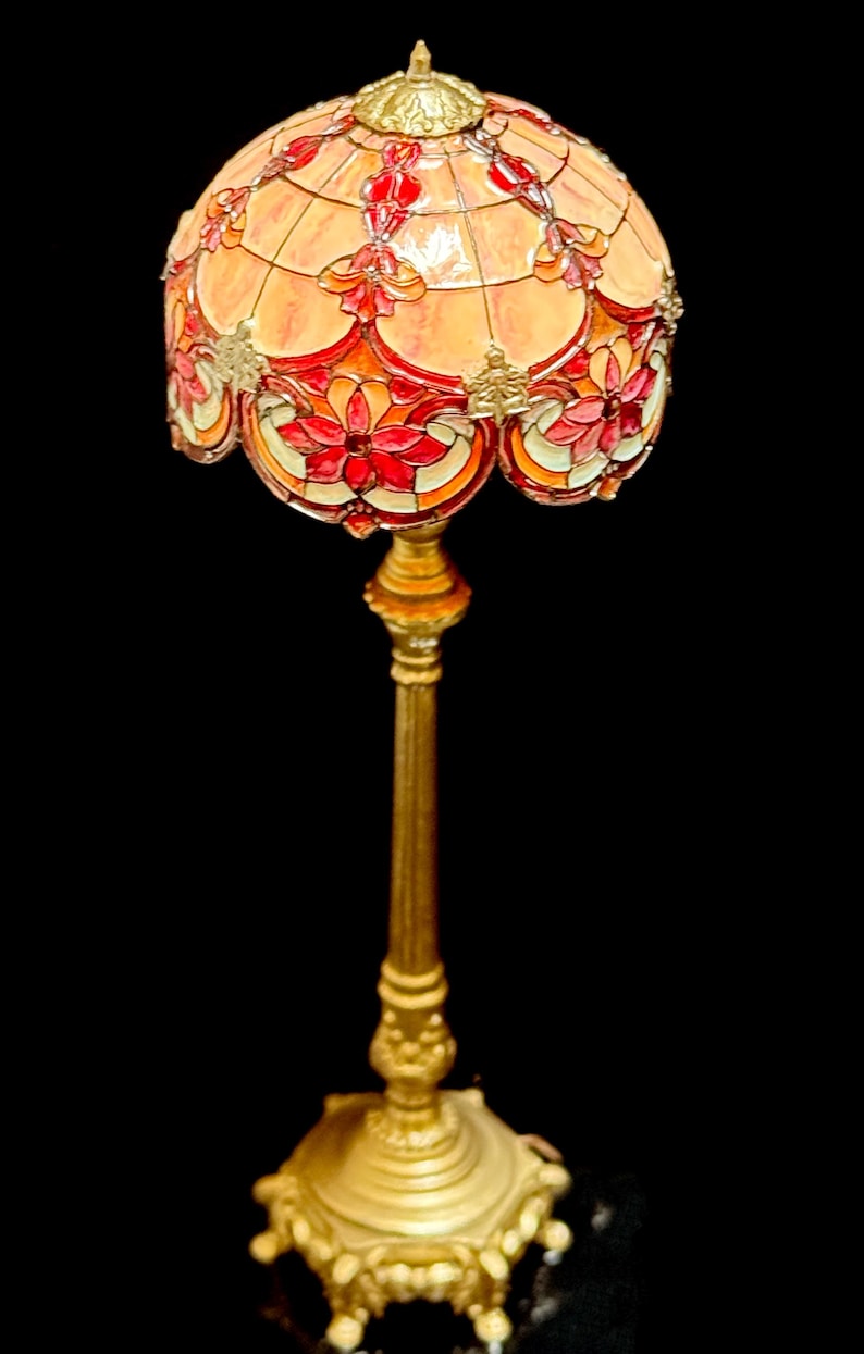 May include: A decorative floor lamp with a stained glass shade. The shade features a floral design in shades of red, orange, and yellow. The base and stem are gold-colored, with ornate detailing. The lamp is a vintage style.