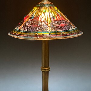 Op de afbeelding: Een miniatuur Tiffany-lamp met een libellenmotief. De lampenkap is gemaakt van kleurrijk glas en heeft een gouden voet.