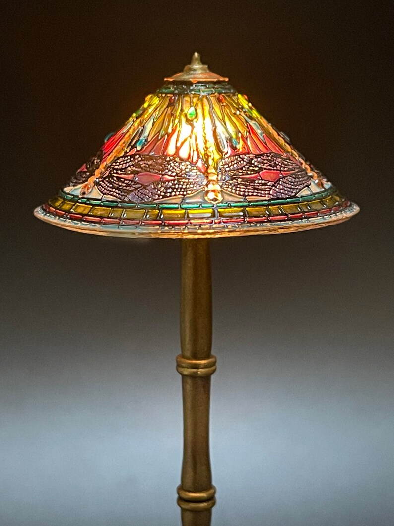 Op de afbeelding: Een miniatuur Tiffany-lamp met een libellenmotief. De lampenkap is gemaakt van kleurrijk glas en heeft een gouden voet.