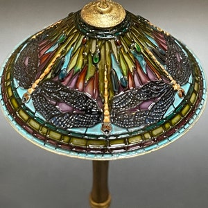 Op de afbeelding: Een lampenkap van gekleurd glas met een libellenmotief in tinten blauw, groen, paars en goud. De lampenkap is gemonteerd op een gouden metalen voet.