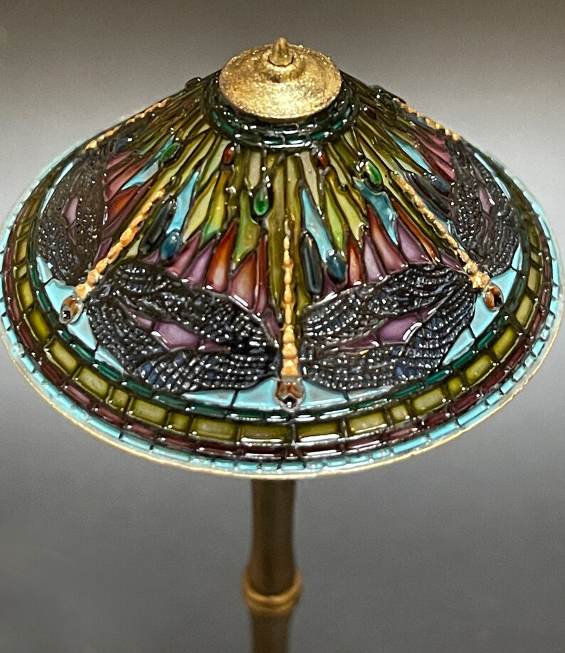 Op de afbeelding: Een lampenkap van gekleurd glas met een libellenmotief in tinten blauw, groen, paars en goud. De lampenkap is gemonteerd op een gouden metalen voet.