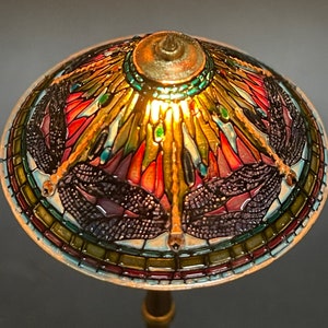 Op de afbeelding: Een lampenkap van gekleurd glas met een libellenmotief. De kap is gemaakt van gekleurde glasstukken in tinten rood, groen, blauw en goud. De lampenkap wordt van binnenuit verlicht en geeft een warm licht.