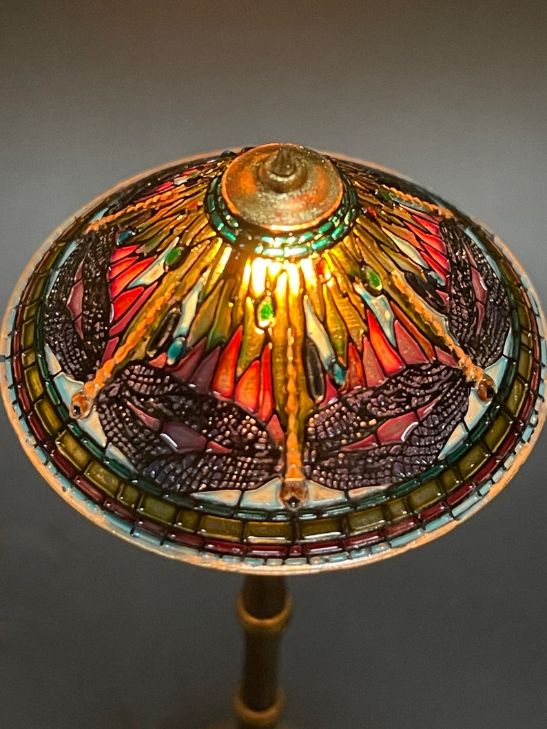 Op de afbeelding: Een lampenkap van gekleurd glas met een libellenmotief. De kap is gemaakt van gekleurde glasstukken in tinten rood, groen, blauw en goud. De lampenkap wordt van binnenuit verlicht en geeft een warm licht.