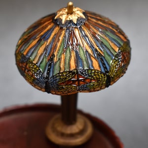 Pode incluir: Uma l&acirc;mpada de vidro Tiffany em miniatura com um design de lib&eacute;lula. O abajur &eacute; feito de pe&ccedil;as de vidro coloridas em tons de azul, verde, laranja e dourado. A base &eacute; de madeira marrom escura com detalhes dourados.
