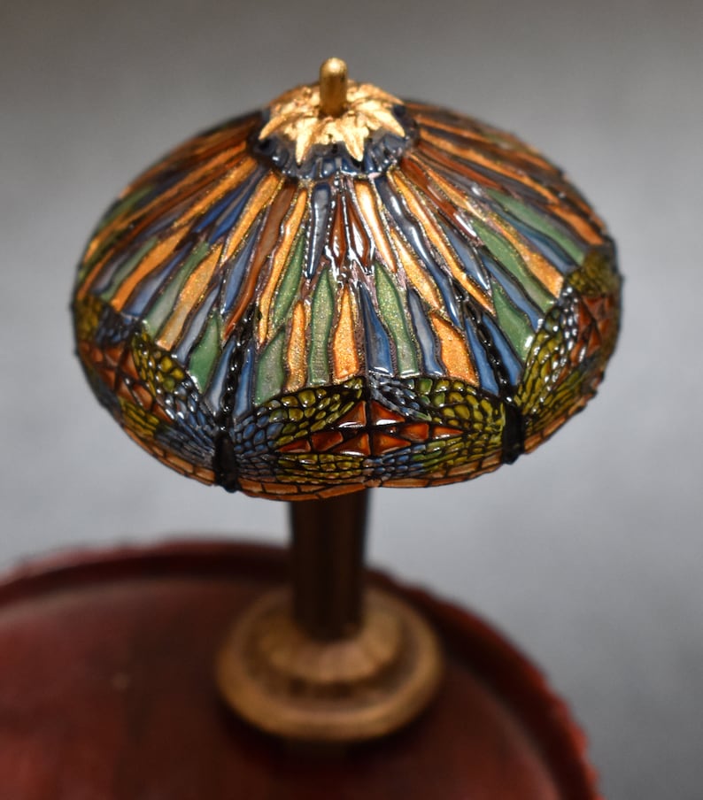 Pode incluir: Uma l&acirc;mpada de vidro Tiffany em miniatura com um design de lib&eacute;lula. O abajur &eacute; feito de pe&ccedil;as de vidro coloridas em tons de azul, verde, laranja e dourado. A base &eacute; de madeira marrom escura com detalhes dourados.