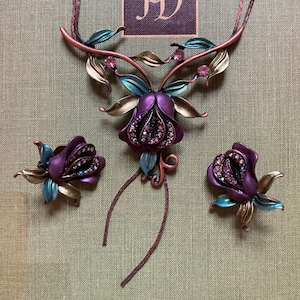 May include: A set of purple and teal floral jewelry with a long necklace and two matching earrings. The necklace features a large flower with a detailed design and a long dangling chain. The earrings are smaller versions of the necklace flower.
