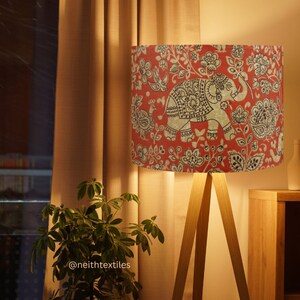 May include: A floor lamp with a cylindrical lampshade featuring a red background with a repeating black and white pattern of elephants, birds, and floral motifs. The lamp has three light-colored wooden legs and emits a warm, soft glow.