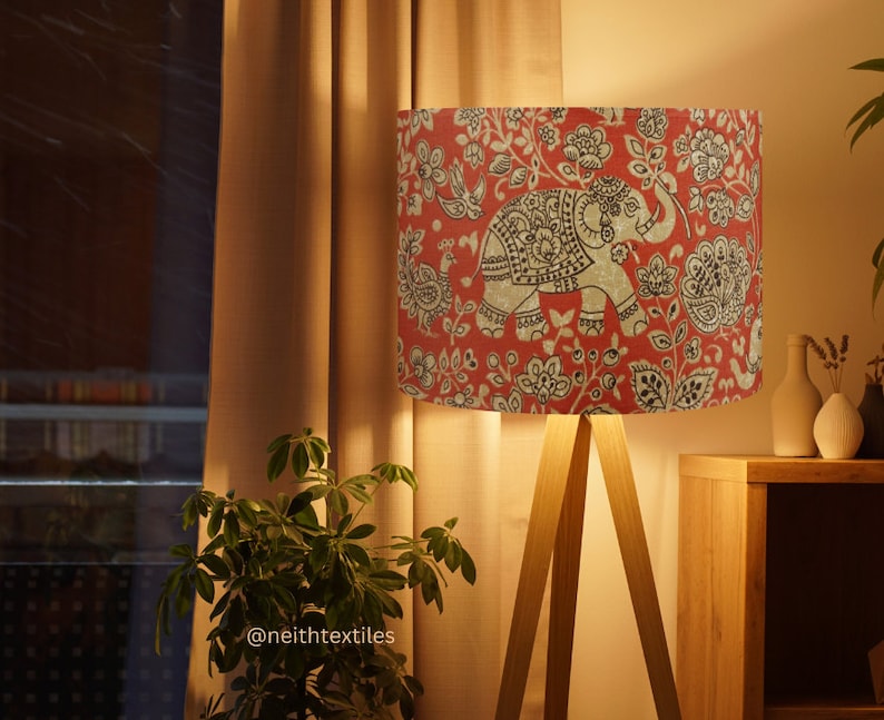 May include: A floor lamp with a cylindrical lampshade featuring a red background with a repeating black and white pattern of elephants, birds, and floral motifs. The lamp has three light-colored wooden legs and emits a warm, soft glow.
