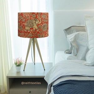 May include: A tripod table lamp featuring a cylindrical shade with a vibrant orange background, decorated with a repeating pattern of beige elephants and intricate floral motifs. The lamp stands on three slender, light-colored wooden legs.