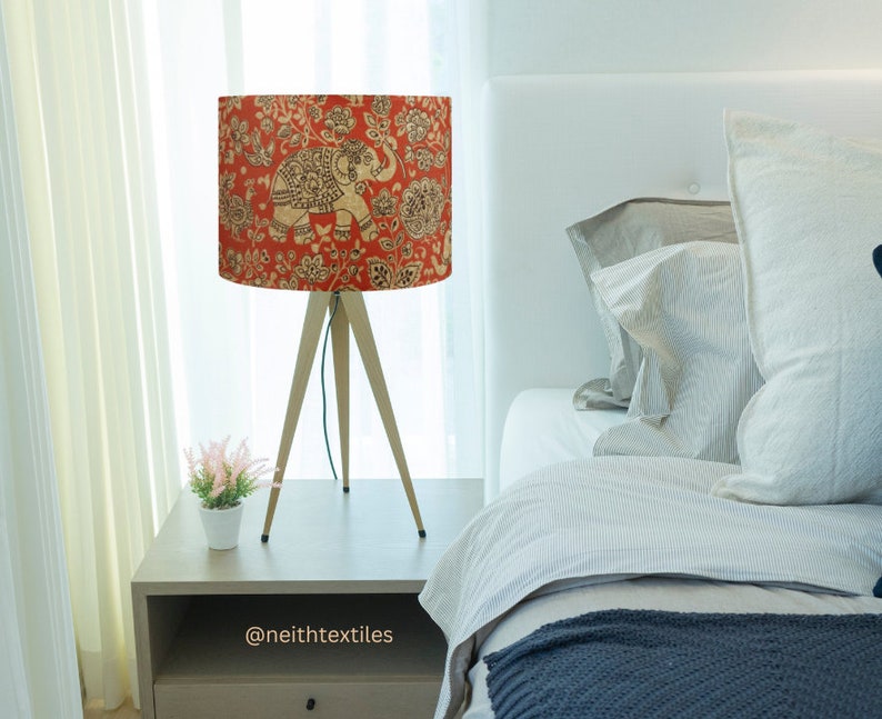 May include: A tripod table lamp featuring a cylindrical shade with a vibrant orange background, decorated with a repeating pattern of beige elephants and intricate floral motifs. The lamp stands on three slender, light-colored wooden legs.