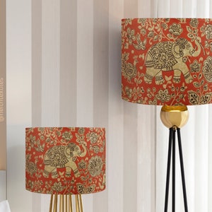 May include: Two cylindrical lamp shades featuring a vibrant red background with a repeating pattern of gold-toned elephants, birds, and floral motifs. The shades are designed for use with lamps and display the text @neithtextiles vertically.