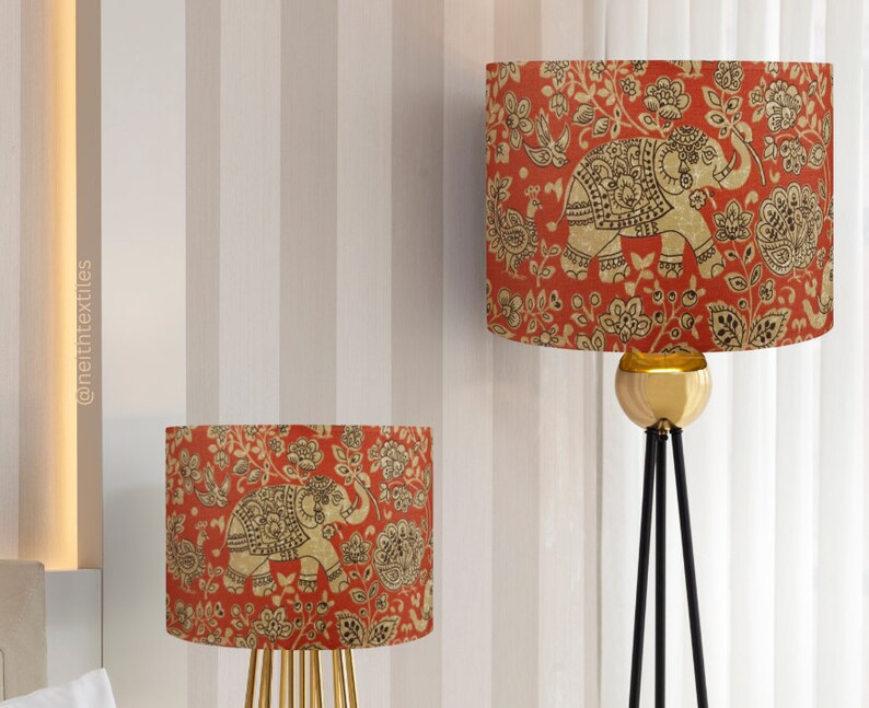 May include: Two cylindrical lamp shades featuring a vibrant red background with a repeating pattern of gold-toned elephants, birds, and floral motifs. The shades are designed for use with lamps and display the text @neithtextiles vertically.