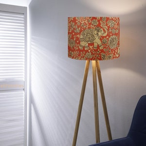 May include: A floor lamp with a cylindrical shade featuring a red background with a gold-toned elephant and floral pattern. The lamp has three light-colored wooden legs arranged in a tripod base. The text @neithtextiles is visible on the left.
