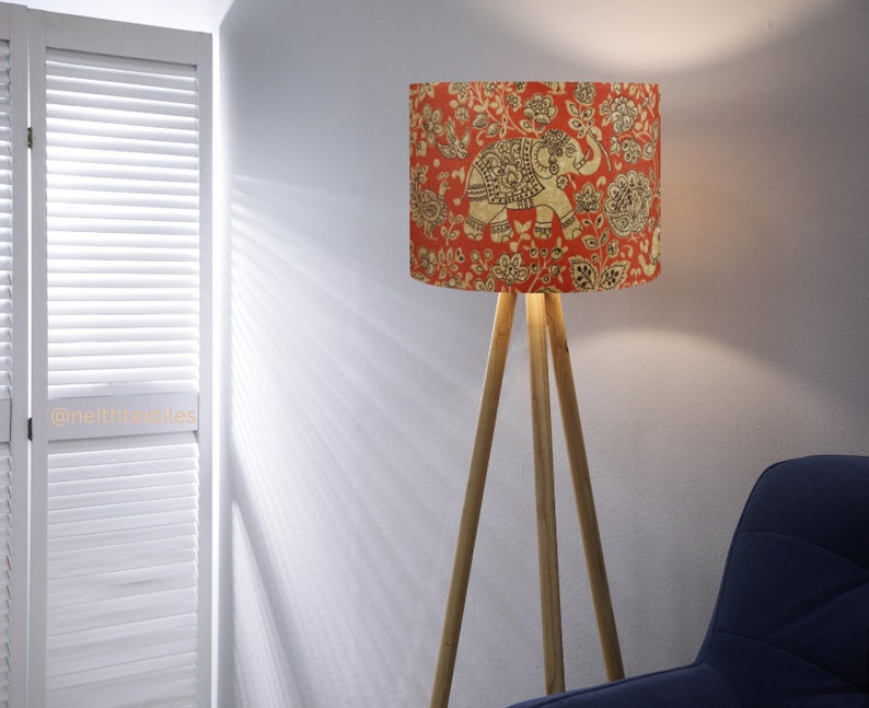 May include: A floor lamp with a cylindrical shade featuring a red background with a gold-toned elephant and floral pattern. The lamp has three light-colored wooden legs arranged in a tripod base. The text @neithtextiles is visible on the left.