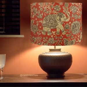 May include: A table lamp with a dark brown, rounded base and a cylindrical lampshade featuring a red background with a black and white pattern of elephants, birds, and floral motifs. The text @neithtextiles is visible on the surface.