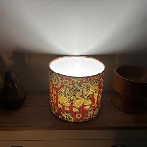 May include: A cylindrical lamp shade featuring a vibrant red fabric background with a repeating gold and black elephant pattern and floral motifs. The shade is illuminated from within, casting a bright white light upward against the wall.