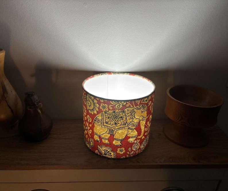 May include: A cylindrical lamp shade featuring a vibrant red fabric background with a repeating gold and black elephant pattern and floral motifs. The shade is illuminated from within, casting a bright white light upward against the wall.