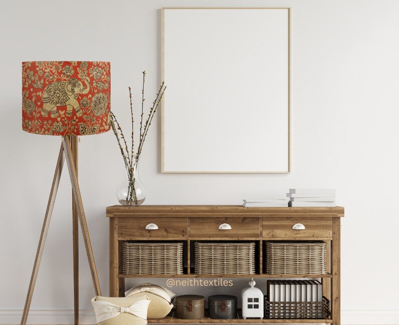 May include: A wooden tripod floor lamp with a red lampshade featuring a gold elephant and floral pattern, a large empty rectangular picture frame, and a wooden console table with three drawers, three wicker baskets, and decor items.