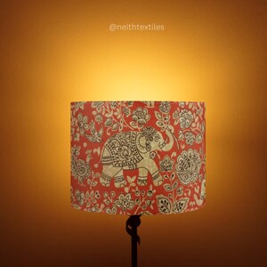 May include: A cylindrical lampshade featuring a red background with a repeating pattern of beige elephants, birds, and floral motifs. The text @neithtextiles is printed in small white letters above the lampshade on the wall.