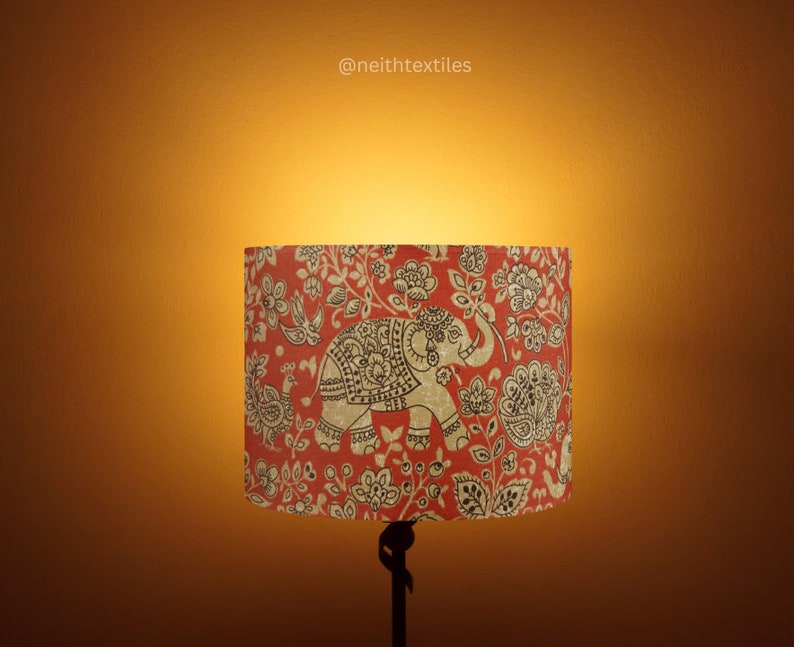 May include: A cylindrical lampshade featuring a red background with a repeating pattern of beige elephants, birds, and floral motifs. The text @neithtextiles is printed in small white letters above the lampshade on the wall.
