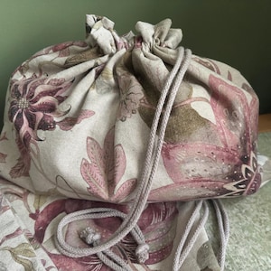 May include: A beige fabric drawstring bag with a floral design in pink, brown, and green. The bag has a drawstring closure. The floral pattern features large, stylized flowers and leaves. The bag is made of fabric.