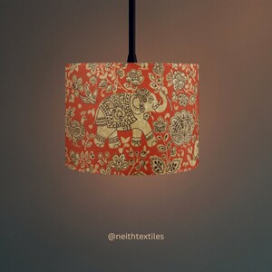May include: A cylindrical lampshade featuring a vibrant orange background with a repeating pattern of dark brown elephants, birds, and floral motifs. The design includes intricate decorative details on the animals. The text @neithtextiles is visible.