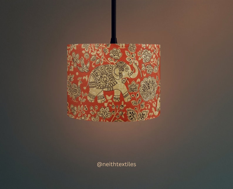 May include: A cylindrical lampshade featuring a vibrant orange background with a repeating pattern of dark brown elephants, birds, and floral motifs. The design includes intricate decorative details on the animals. The text @neithtextiles is visible.