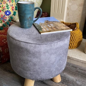 May include: A grey, round, upholstered footstool with wooden legs. The footstool is sitting on a wooden floor and has a book and a mug on top of it.