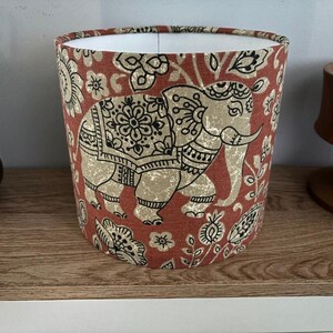 May include: A cylindrical lampshade featuring a rust-orange fabric background with a repeating pattern of beige elephants and black floral motifs. The interior of the lampshade is white, and the exterior displays detailed black line work on the elephants.