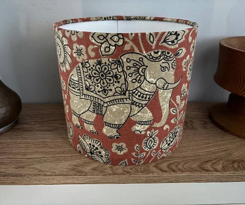 May include: A cylindrical lampshade featuring a rust-orange fabric background with a repeating pattern of beige elephants and black floral motifs. The interior of the lampshade is white, and the exterior displays detailed black line work on the elephants.