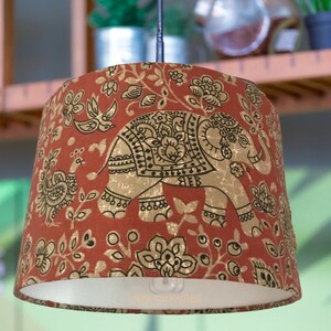 May include: A drum-shaped pendant lampshade featuring a burnt orange fabric base with a repeating gold and black print of elephants, peacocks, and floral motifs. The interior is white, and the lower edge displays the text nerthtextiles.