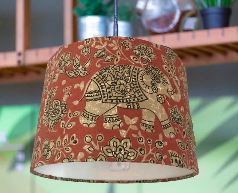 May include: A drum-shaped pendant lampshade featuring a burnt orange fabric base with a repeating gold and black print of elephants, peacocks, and floral motifs. The interior is white, and the lower edge displays the text nerthtextiles.