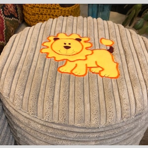 May include: A gray corduroy round pouf with a yellow lion applique. The lion has orange eyes and a black nose.