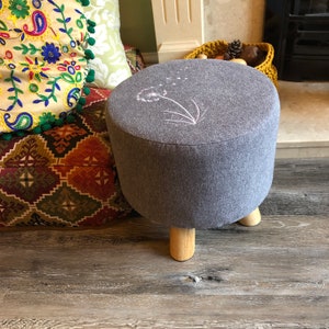 May include: A gray round ottoman with wooden legs and a white embroidered dandelion design.
