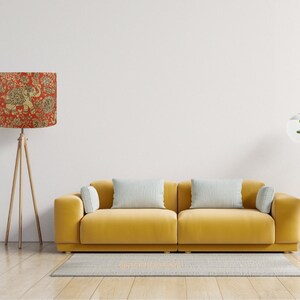 May include: A floor lamp with a cylindrical shade featuring a red background and a gold elephant and floral pattern, standing on a wooden tripod base next to a yellow sofa with two light gray rectangular pillows and two smaller side cushions.