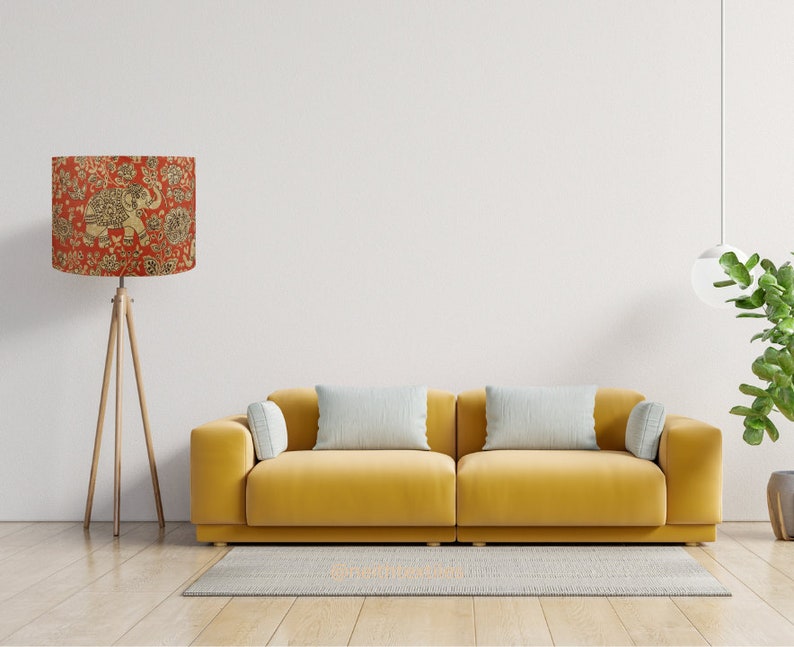 May include: A floor lamp with a cylindrical shade featuring a red background and a gold elephant and floral pattern, standing on a wooden tripod base next to a yellow sofa with two light gray rectangular pillows and two smaller side cushions.
