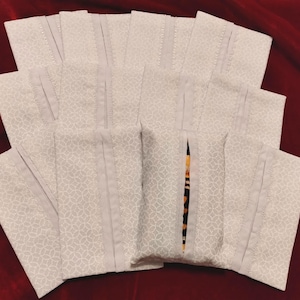 May include: A collection of light gray fabric pouches with a subtle floral pattern. Each pouch has a vertical seam down the center and a contrasting light gray border. The pouches are arranged on a deep red velvet surface.