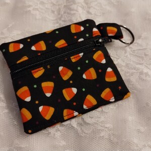 May include: A small square fabric coin pouch featuring a black background with a repeating pattern of orange, yellow, and white candy corn shapes and small green dots. The pouch has a black zipper closure and a silver metal keyring attached to the side.