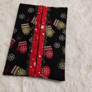 May include: A rectangular fabric tissue holder featuring a black background with a repeating pattern of white snowflakes and red and green mittens. A vertical red fabric strip with white polka dots runs down the center opening of the holder.